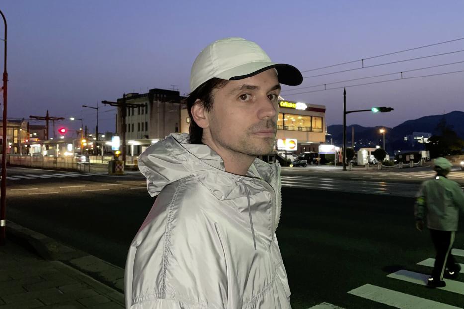 Mann in weißer Kappe und heller Jacke steht an einer nächtlichen Kreuzung mit Ampeln und Bergen im Hintergrund.