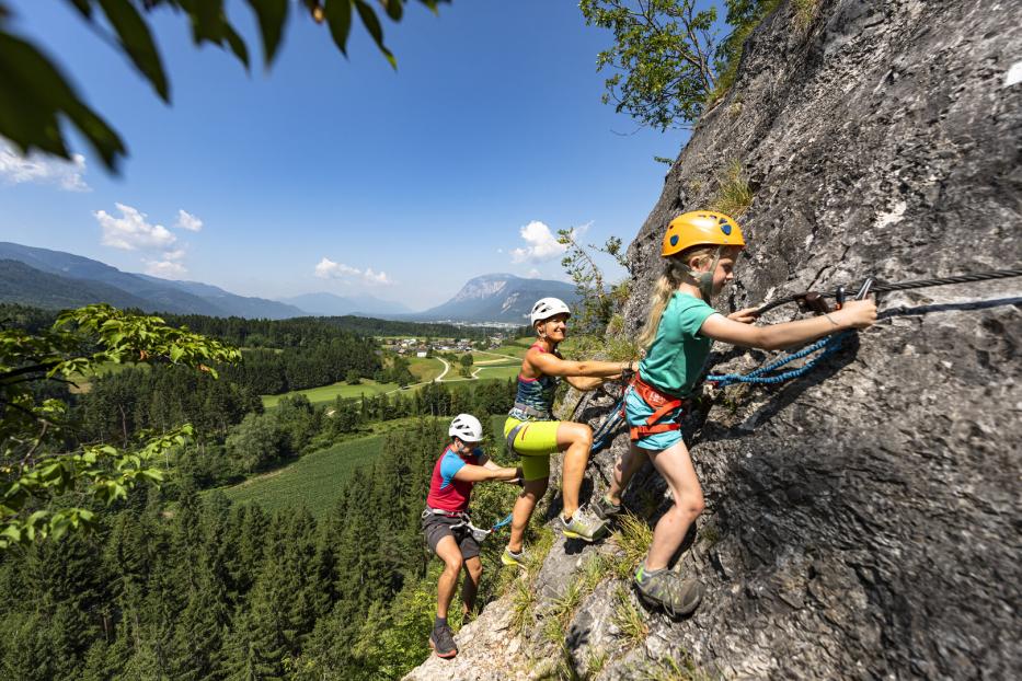Eine Familie klettert gesichert an einem Felsen mit Blick auf eine grüne Berglandschaft.