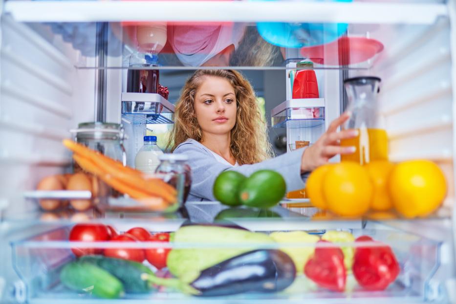 Eine junge Frau mit lockigem Haar greift aus der Perspektive des Kühlschrankinneren nach einem Glas Saft zwischen frischem Obst und Gemüse.