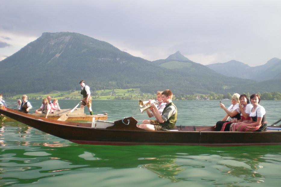 Mehrere Menschen in Tracht fahren und musizieren in Holzbooten auf einem See vor Bergen.