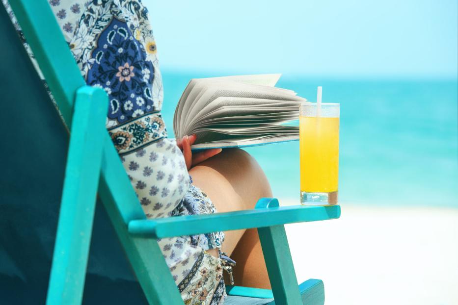 Woman sitting in deck chair at beach reading book on sunny day.