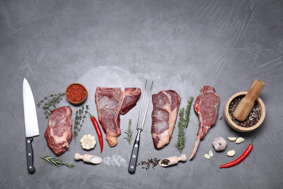 Vier rohe Steaks mit Messern, Kräutern, Knoblauch, Chilischoten und Gewürzen auf grauem Hintergrund.
