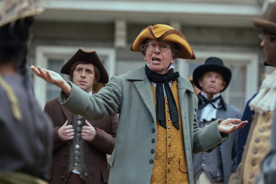 Ein Mann (Larry David) mit Brille und in historischer Kleidung mit Dreispitz gestikuliert lebhaft vor einer Gruppe weiterer Personen in zeitgenössischen Kostümen.