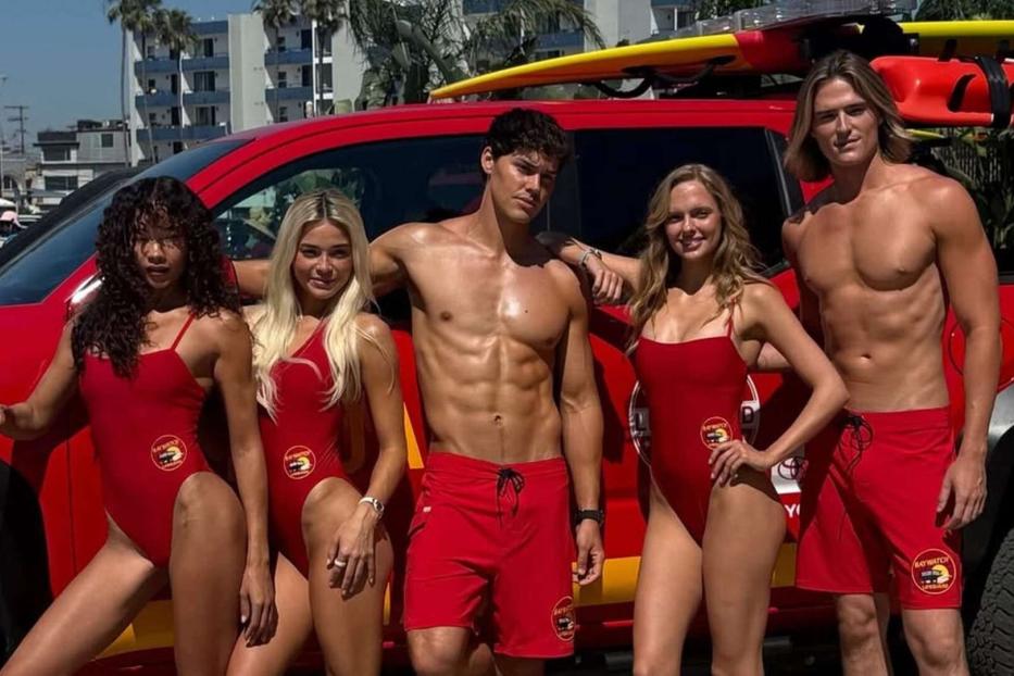 Fünf Rettungsschwimmer in roten Badeanzügen und Shorts posieren vor einem roten Lifeguard-Pick-up mit Surfbrettern auf dem Dach.