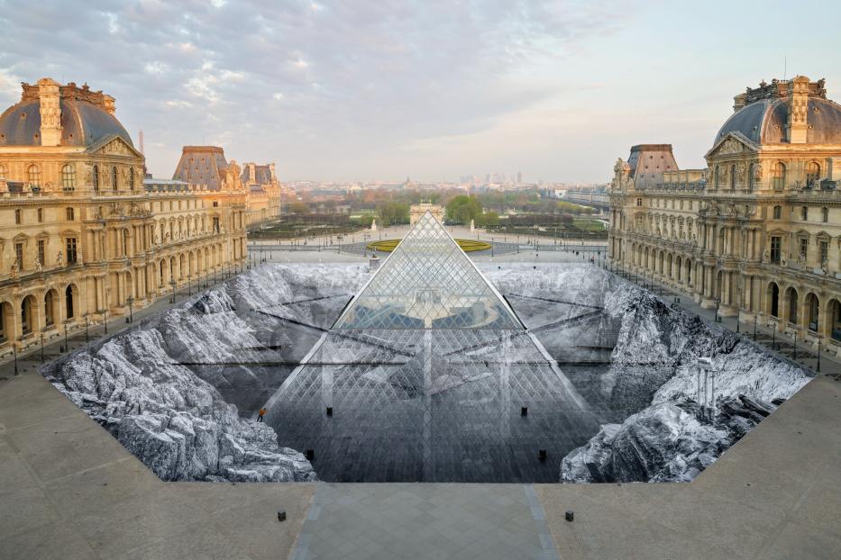 Die Glaspyramide des Louvre in Paris ist von einer großflächigen, schwarz-weißen Bodeninstallation umgeben, die eine felsige Landschaft darstellt.