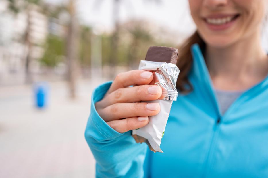 Lächelnde Frau in blauer Jacke hält einen ausgepackten Schokoriegel in der Hand.