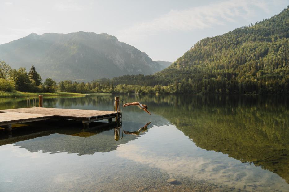 Eine Frau springt von einem Steg in einen stillen Bergsee, umgeben von bewaldeten Hängen und Spiegelungen im Wasser.