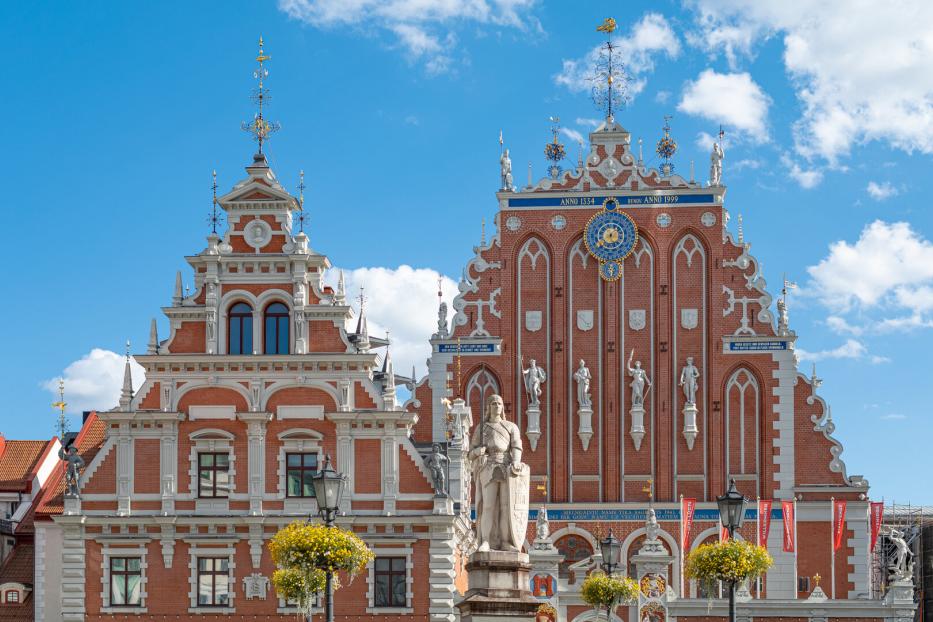 Das Schwarzhäupterhaus in Riga mit seiner verzierten Backsteinfassade und einer davorstehenden Roland-Statue unter blauem Himmel.