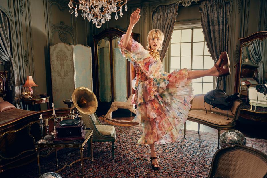 Eine Frau in luftiger Blumenkleidung tanzt mit erhobenem Bein in einem opulent eingerichteten Schlafzimmer.