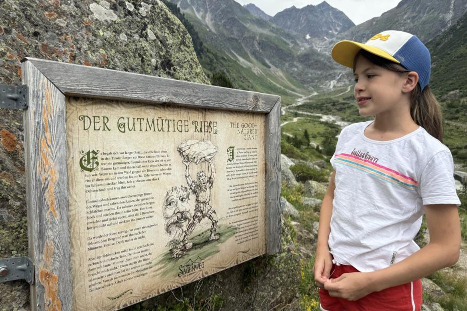Ein Mädchen liest vor einer Bergkulisse eine Informationstafel über den „Gutmütigen Riesen“ am Pitztaler Sagenweg.