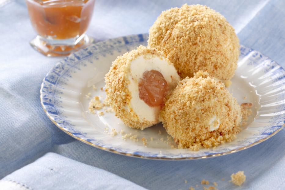 Marillenknödel mit Bröseln auf einem Teller, daneben ein kleines Glas mit Aprikosenkonfitüre auf einem blauen Tuch.