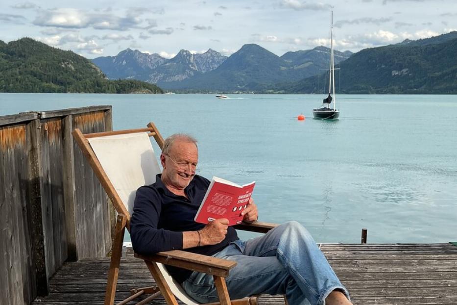 Ein Mann liest entspannt auf einer Holzterrasse am See, mit Segelboot und Bergen im Hintergrund.