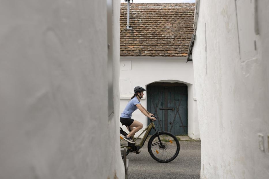 Fahrradfahrerin mit Helm fährt durch eine enge Gasse vor einem weißen Haus mit blauem Tor.