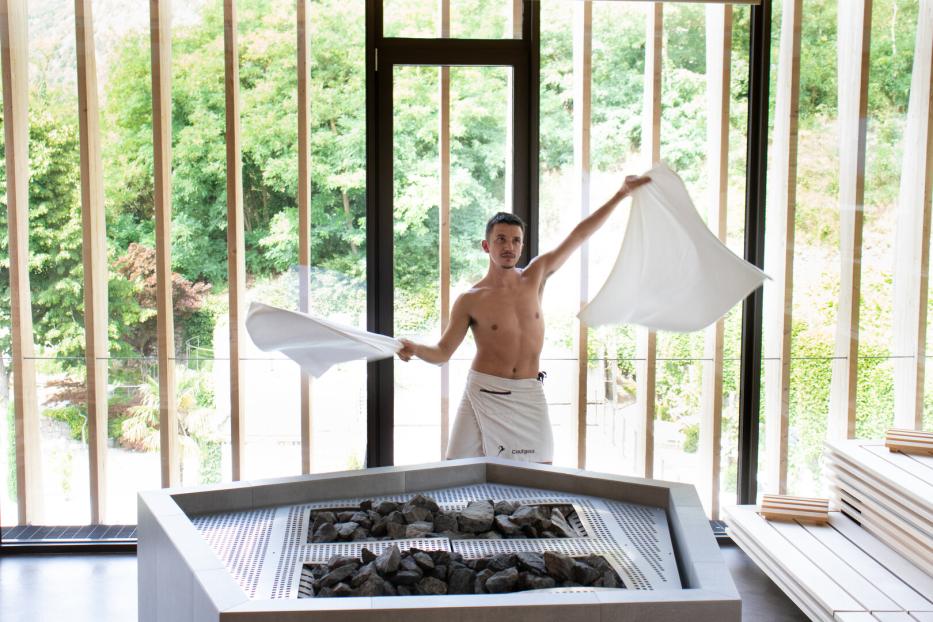 Ein Saunameister schwingt in einer modernen Sauna mit Glasfront zwei weiße Handtücher vor einem Saunaofen.