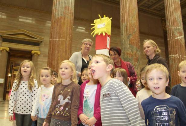 Eine Gruppe Kinder und Erwachsene steht in einem Raum mit Säulen und singt.