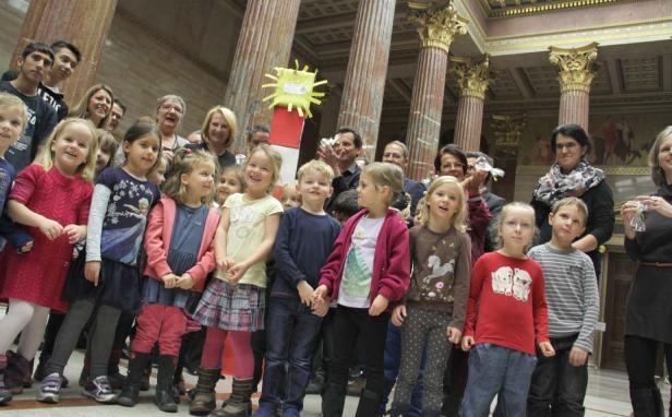 Eine Gruppe von Kindern und Erwachsenen steht in einem prunkvollen Gebäude mit Säulen.