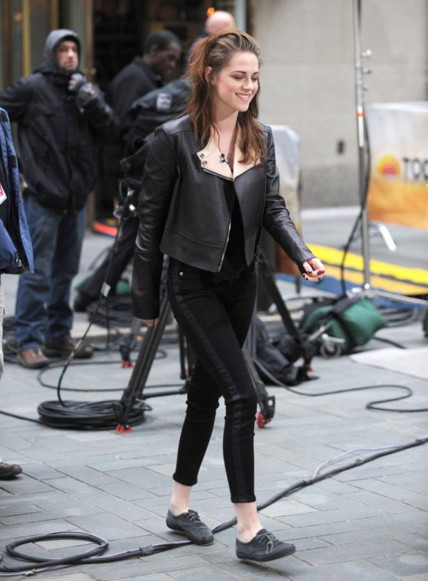 Kristen Stewart lächelt in einer schwarzen Lederjacke und schwarzen Jeans auf einem Filmset.