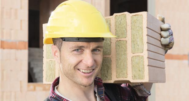 Ein Bauarbeiter mit gelbem Helm trägt einen isolierten Ziegelstein auf der Schulter.