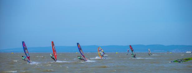 Mehrere Windsurfer rasen über einen See vor einer Hügellandschaft.