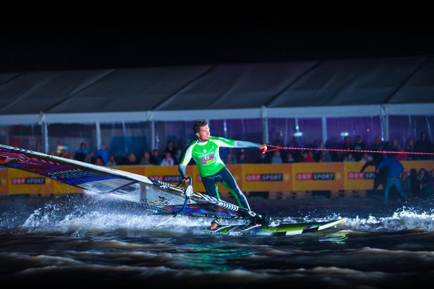 Ein Windsurfer auf dem Wasser vor einer Zuschauermenge bei Nacht.