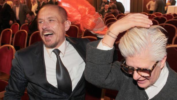 Ein Mann hält eine orangefarbene Perücke über den Kopf einer Frau mit Brille.