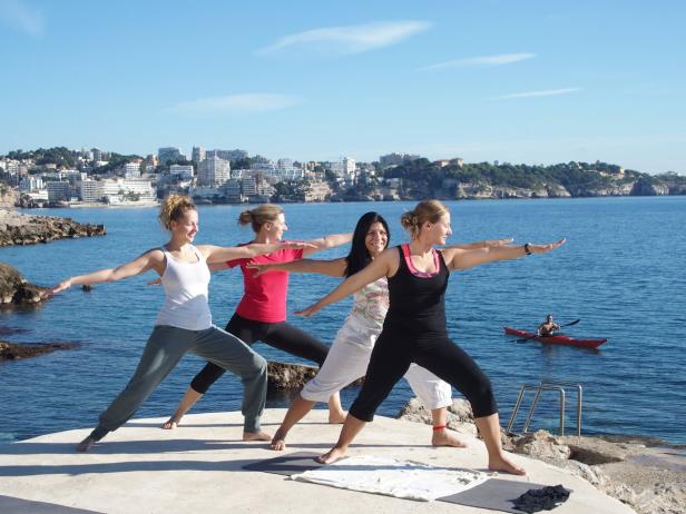 Eine Gruppe von Frauen macht Yoga am Meer, während im Hintergrund ein Mann in einem Kajak paddelt.