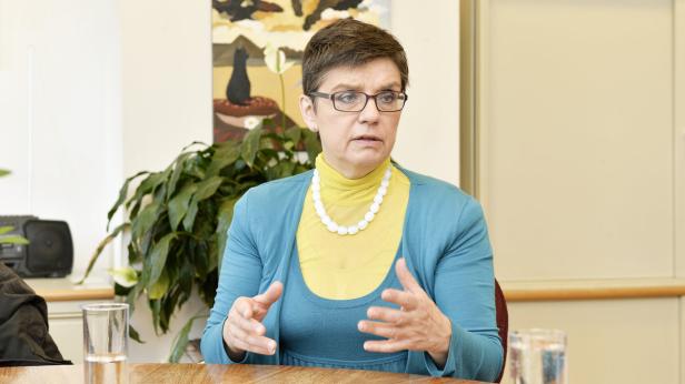 Eine Frau mit Brille und Halskette sitzt an einem Tisch und gestikuliert.