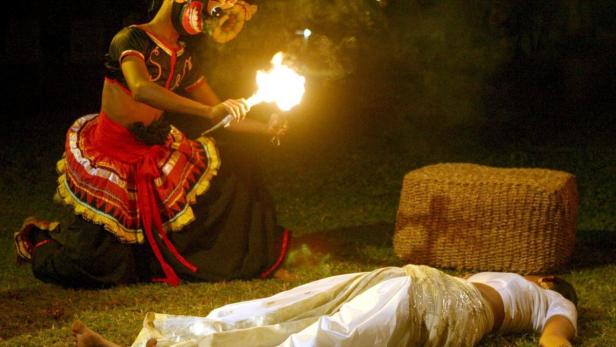 Eine maskierte Tänzerin mit einer Fackel führt einen traditionellen Tanz vor einer liegenden Person auf.