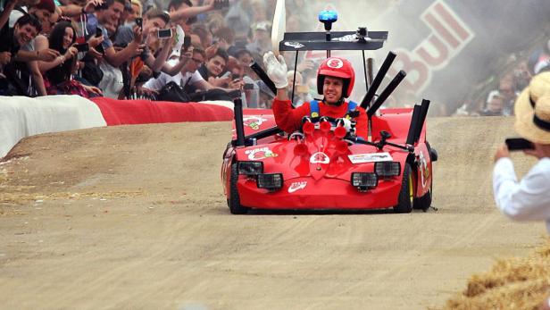 Ein Mann in einem roten, selbstgebauten Seifenkistenwagen winkt dem Publikum zu.