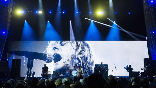 Eine Rockband tritt vor einem großen Bildschirm mit dem Gesicht eines Sängers auf.