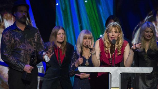 Courtney Love spricht bei einer Veranstaltung am Rednerpult, umgeben von anderen Personen.