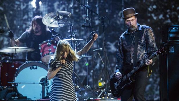 Eine Sängerin mit gestreiftem Kleid singt, während ein Bassist und ein Schlagzeuger spielen.