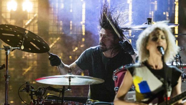 Dave Grohl spielt Schlagzeug, während eine Sängerin im Vordergrund singt.