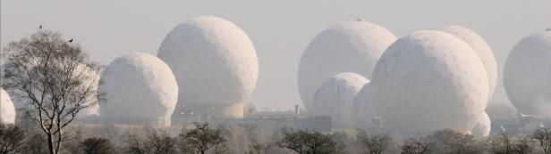 Mehrere weiße Radarkuppeln erheben sich über eine bewaldete Landschaft.