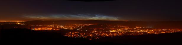 Blick auf eine Stadt bei Nacht mit leuchtenden Nachtwolken am Himmel.