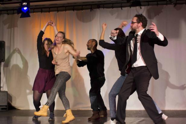 Eine Gruppe von fünf Personen tanzt auf einer Bühne vor einem weißen Vorhang.