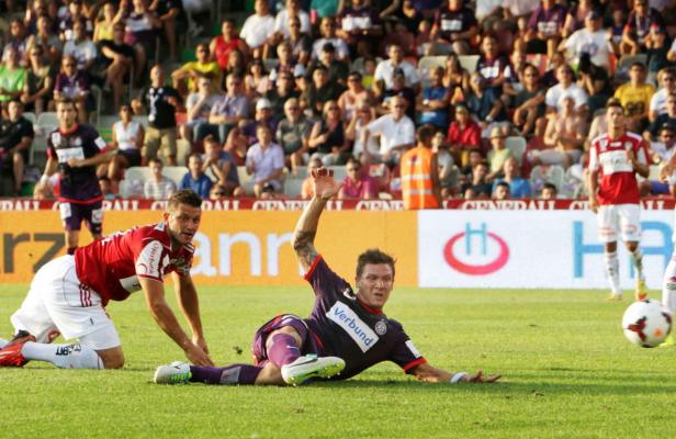Zwei Fußballspieler kämpfen auf dem Rasen um den Ball, während Zuschauer im Hintergrund sitzen.