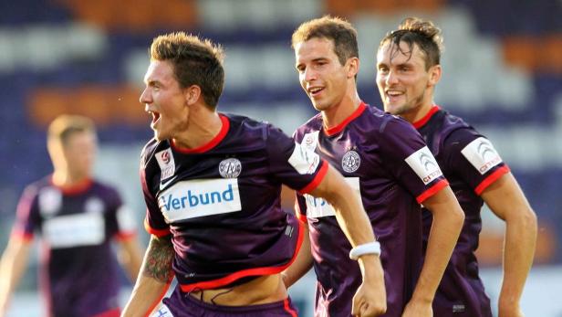 Drei Fußballspieler in lila Trikots jubeln auf dem Spielfeld.