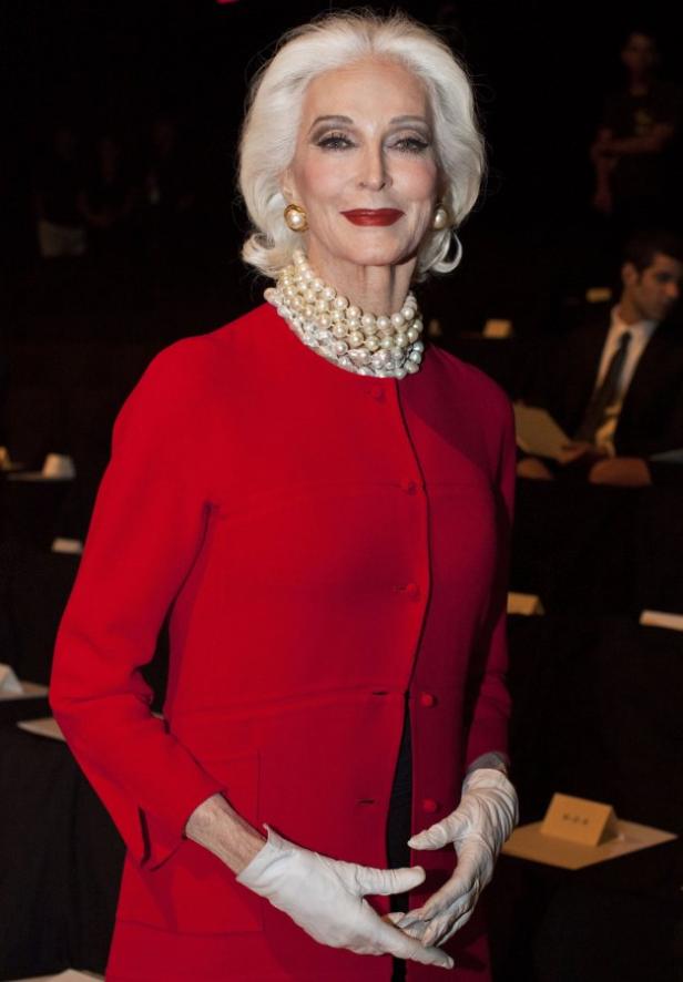 Eine ältere Frau mit weißem Haar, rotem Blazer, Perlenkette und weißen Handschuhen.