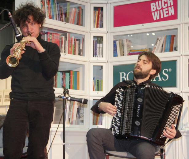 Zwei Musiker, einer mit Saxophon, der andere mit Akkordeon, treten vor einem Bücherregal auf.