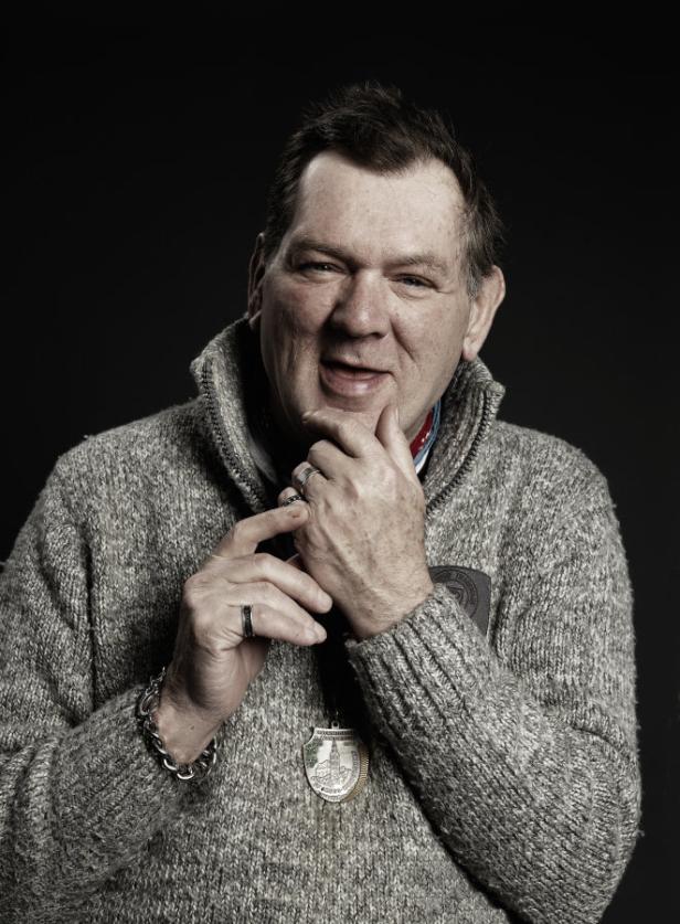 Der Gugginger Künstler Johann Garber (Braschler/Fischer) Ein Mann mit Medaille und grauem Strickpullover vor dunklem Hintergrund.