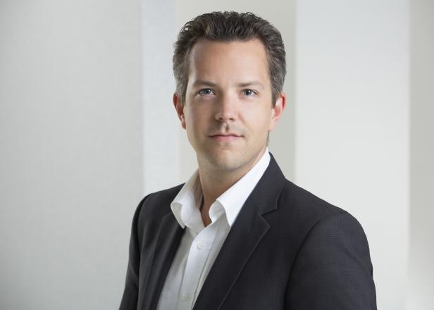Ein Mann mit dunklen Haaren trägt ein schwarzes Sakko über einem weißen Hemd.