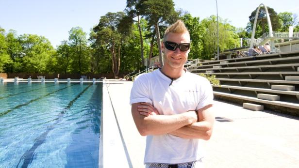 Ein Mann mit Sonnenbrille steht vor einem Schwimmbad mit Tribüne.