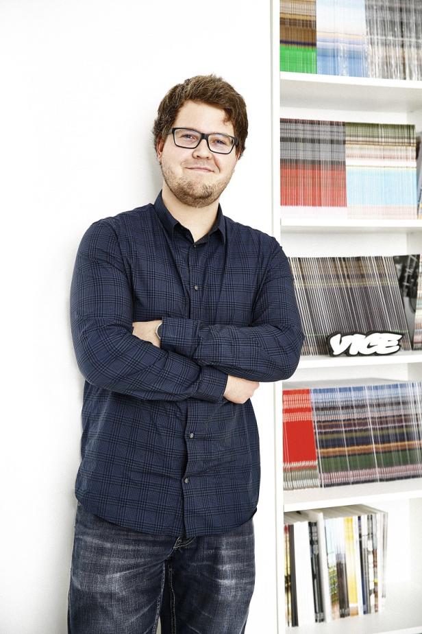 Ein Mann mit Brille und kariertem Hemd steht vor einem Bücherregal mit „Vice“-Magazinen.