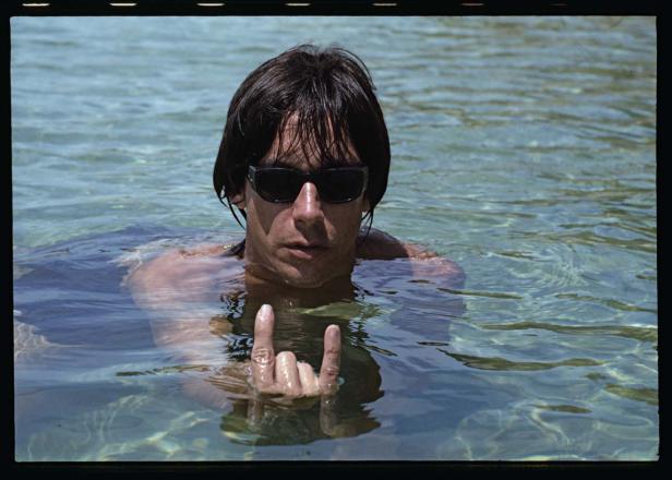 Ein Mann mit Sonnenbrille steht im Wasser und zeigt das Handzeichen „Pommesgabel“.
