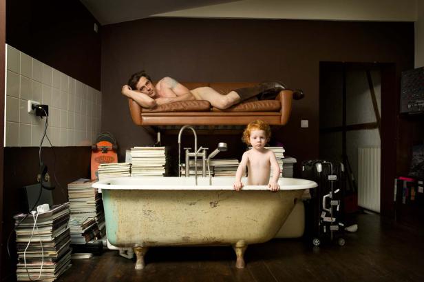 Ein Mann liegt auf einem Sofa über einer Badewanne, in der ein Kind steht.