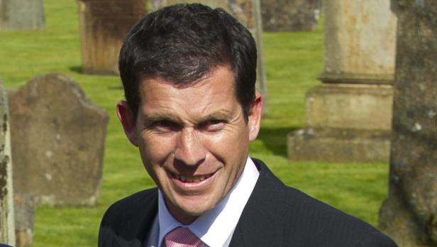 Ein lächelnder Mann in Anzug und Krawatte steht auf einem Friedhof.