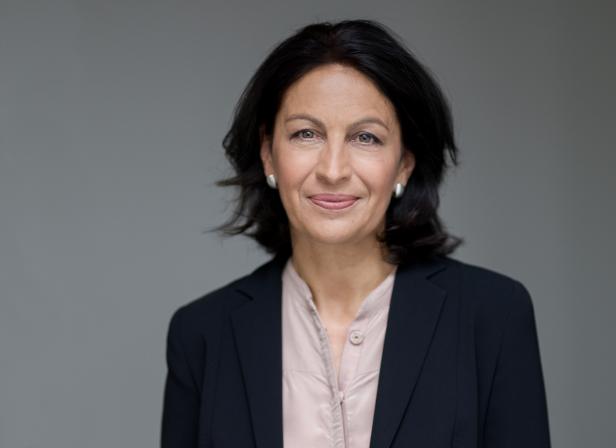 Eine Frau mit dunklen Haaren und einem Blazer lächelt in die Kamera.