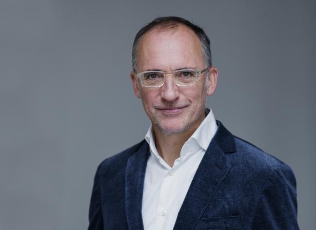 Ein Mann mit Brille und blauem Sakko lächelt in die Kamera.