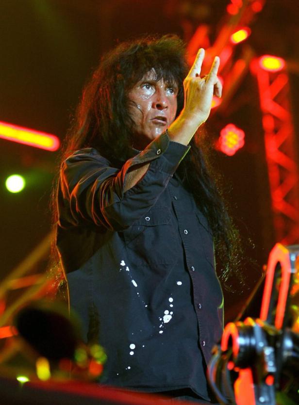 Ein Mann mit langen, dunklen Haaren macht auf einer Bühne das „Pommesgabel“-Zeichen.